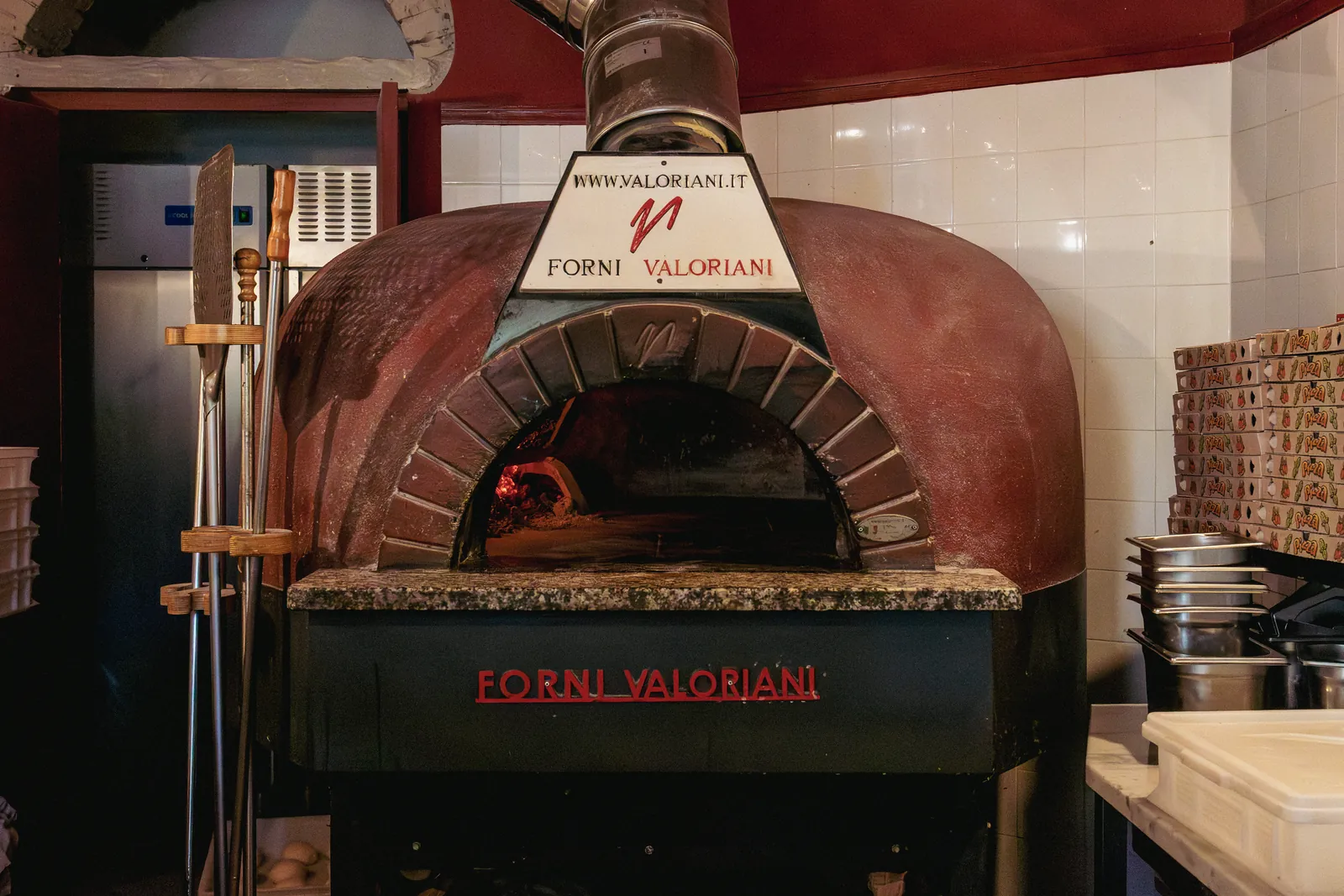 Il forno a legna Valoriani a cupola rossa, acceso, fra le piastrelle bianche della cucina, con le pale appese a sinistra.