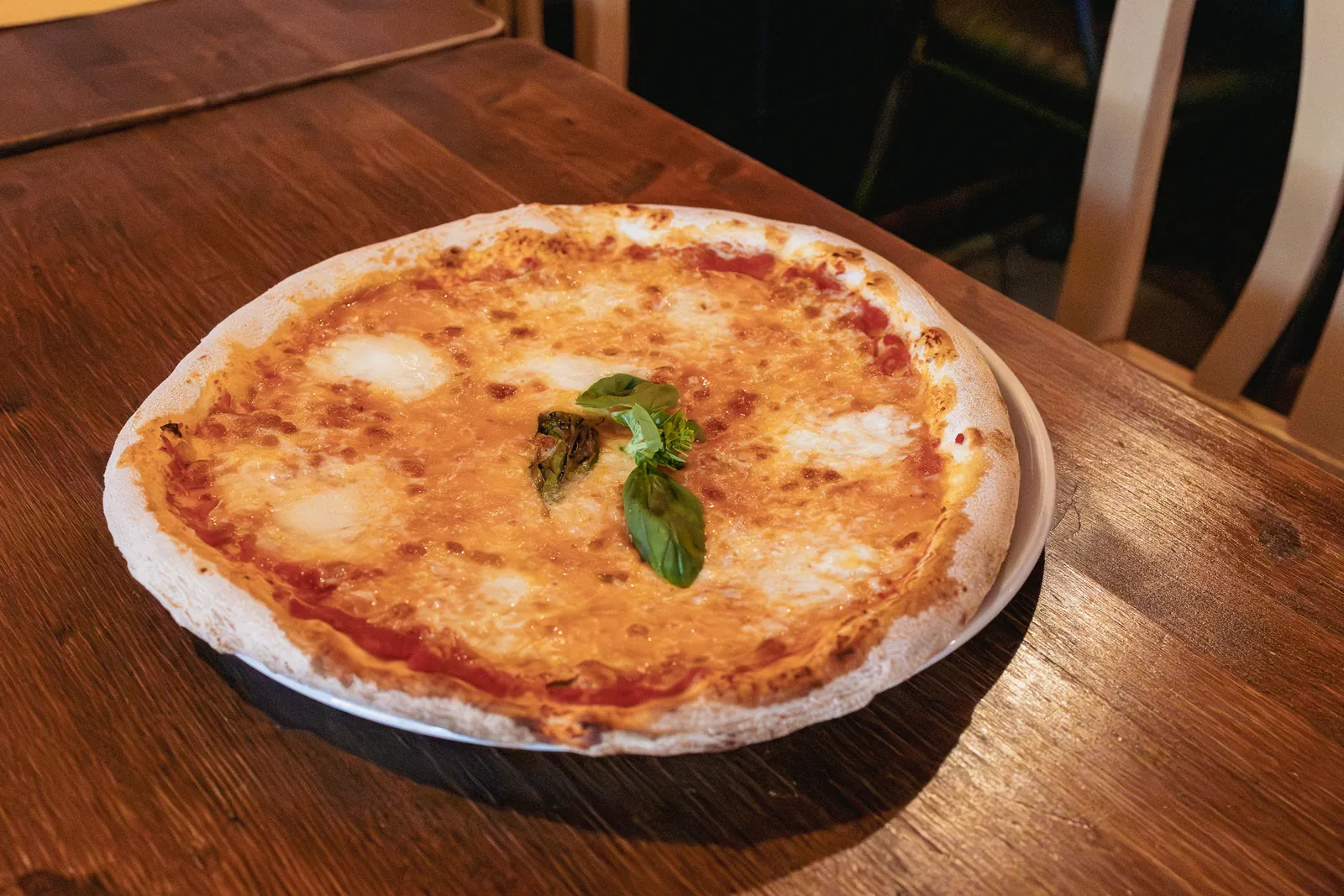 Una pizza Margherita con fiordilatte e foglie di basilico al centro, su un piatto bianco sopra un tavolo di legno.