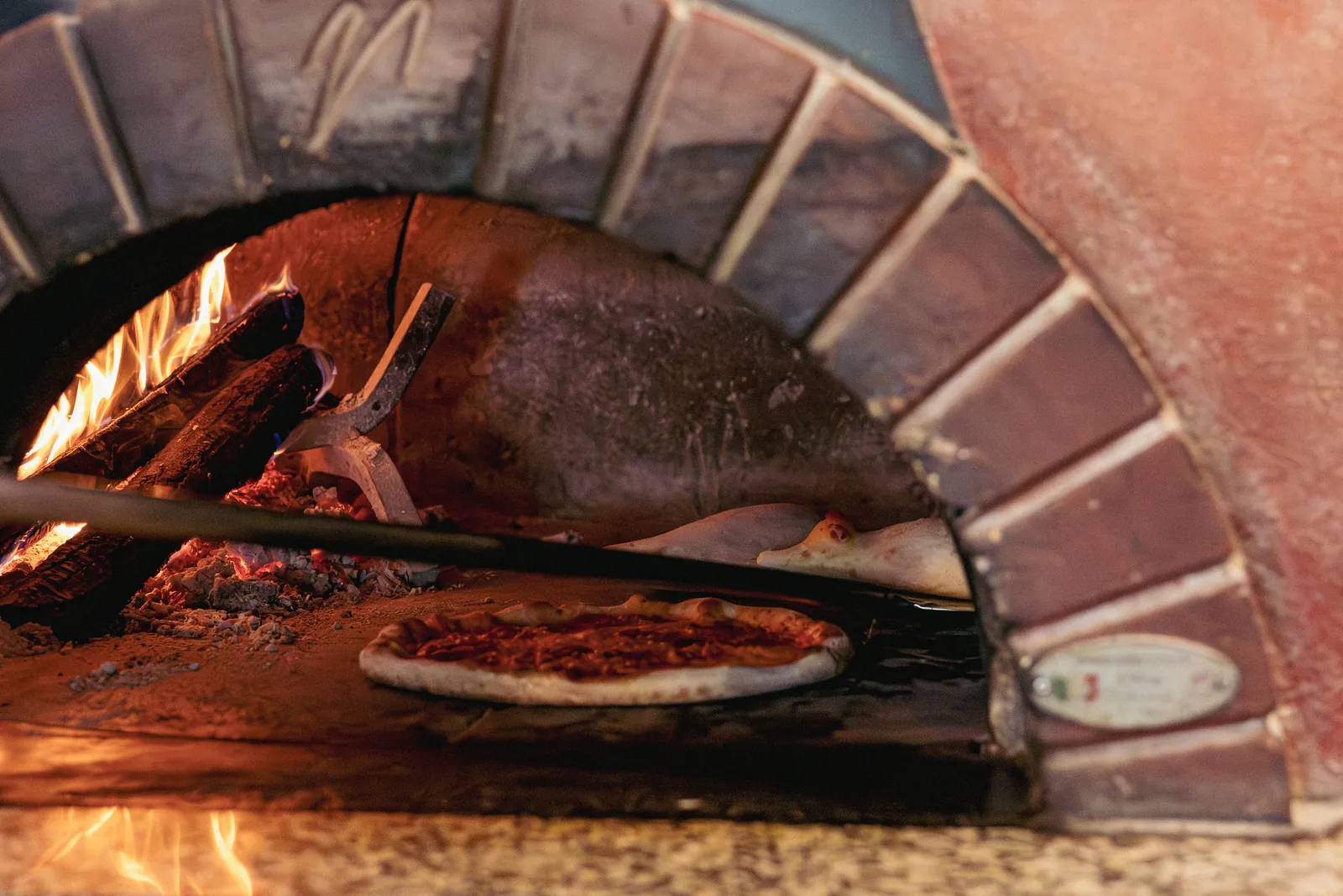 Una pizza sulla pala dentro la bocca del forno, accanto alla legna che arde.