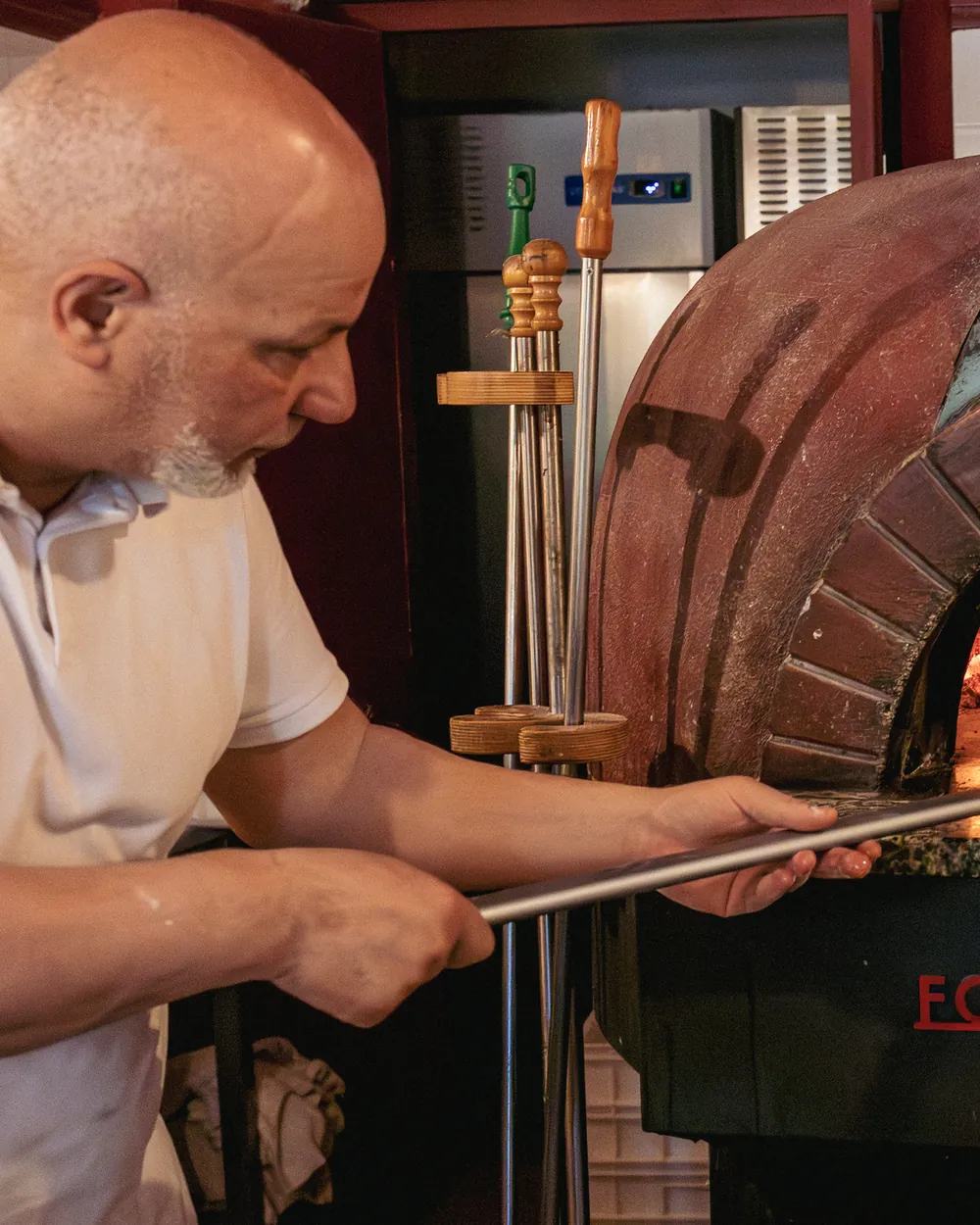 Il pizzaiolo, in maglietta bianca, inforna una pizza con la pala lunga davanti al forno a cupola rossa.