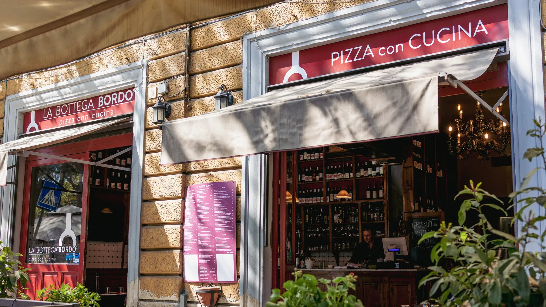 La facciata della Bottega Bordò con le due insegne bordò, La Bottega Bordò pizza con cucina e Pizza con cucina, la tenda, le piante e la vetrina aperta sul banco.
